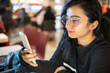 © Bhawna - High angle image of an Asian, Indian beautiful, serene young woman in eyeglasses sitting at a restaurant and busy in using a smartphone.