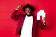 Real estate. Happy Black man celebrating, buying new house, showing paper home model and raising fist up in triumph, standing over red background