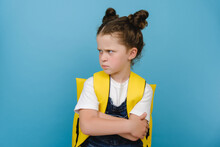 Portrait Of Angry Sad Little Schoolgirl, Crossed Arms Looking To Side, Does Not Want To Go To School, Wears Yellow Backpack, Posing Isolated Over Blue Studio Background. Children's Emotions Concept