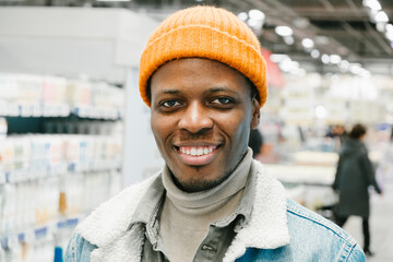 Wall Mural - Cheerful young African-American guy in warm denim jacket stands near refrigerators in modern supermarket department closeup