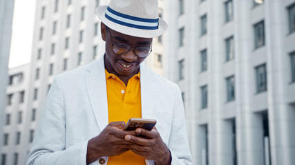 Wall Mural - Smiling African American man in glasses and hat types on smartphone and smiles with happiness