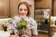© Cavan Images - Smiling woman smells fresh cut lilacs in her kitchen