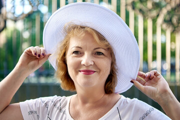 Wall Mural - Senior beautiful woman 54 years old in white hat smiles while walk outside.