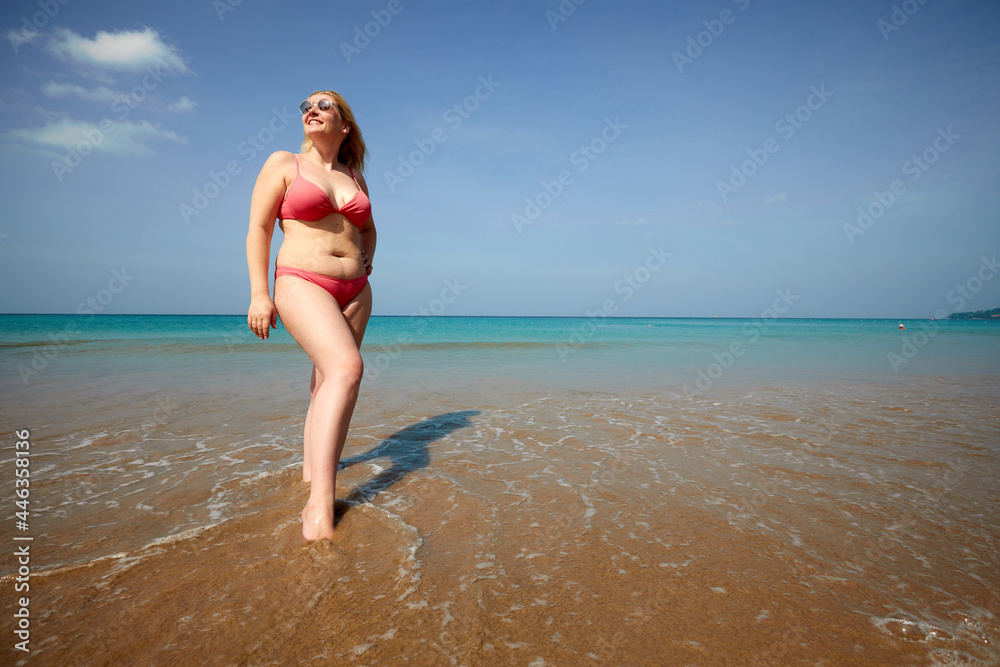 Full body of smiling overweight woman covering face from sunlight while standing on sandy beach near waving sea - obrazy, fototapety, plakaty 