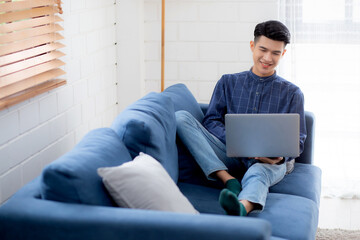 Wall Mural - Young asian businessman smile and work from home with laptop computer online to internet on sofa in living room, freelance man using notebook on couch with comfort, new normal, lifestyle concept.