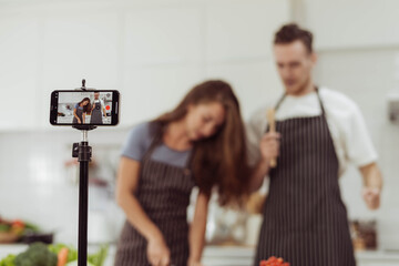 Blogger and Youtuber concept, Happy couple love recording video while cooking together in the kitchen.Focus on smartphone, couple cooking together on live streaming.