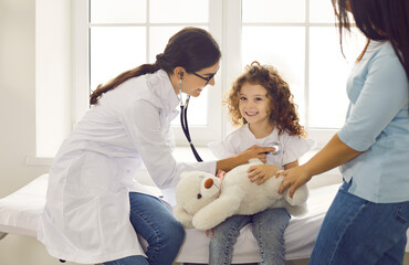 Happy cute child patient with toy seeing family doctor. Female pediatrician with stethoscope checking young patient's heartbeat or doing her lungs exam during medical checkup at hospital or clinic