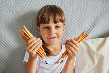 Portrait of charming dark haired girl holding pretzels sticks in hands and looking directly at camera, female child wearing white t shirt, has pigtails, posing at home while sitting on sofa.