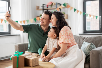 Wall Mural - family, holidays and people concept - portrait of happy mother, father and little son with gifts and smartphone taking selfie on birthday at home party