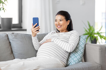 Poster - pregnancy, rest, people and expectation concept - happy smiling pregnant asian woman with smartphone sitting on sofa at home