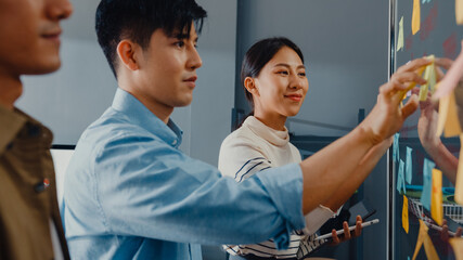 Wall Mural - Asia businesspeople stand behind transparent glass wall listen manager pointing progress work and brainstorm meeting and worker post sticky note on wall at office. Business inspiration, Share ideas.