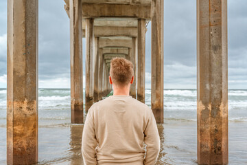 Canvas Print - Young man is standing under pier in La Jolla Shores at the cloudy time. Symmetrical Pier in San Diego, California