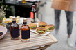 Bottles with liqueur or berry tinctures on a table with burgers, people grilling on a background, close-up on bottles with blank labels for copy paste