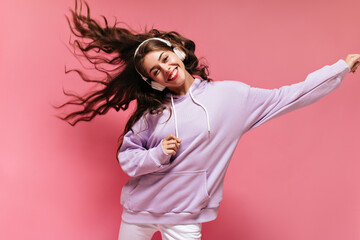 Poster - Pretty woman in oversized purple hoodie dances on isolated pink background. Brunette girl listening to music in headphones.