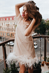 Wall Mural - Pretty woman in white dress moves on balcony. Happy young lady with red lips smiles sincerely and plays hair outside.