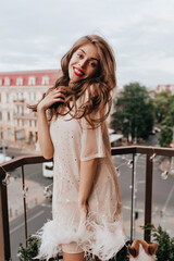 Wall Mural - Cheerful girl with red lips smiles sincerely. Pretty brunette woman in white dress with sparkles poses on balcony in good mood.