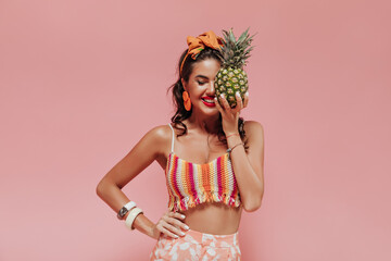 Wall Mural - Optimistic trendy lady with orange bandana and red lips in fashionable summer clothes posing with pineapple and closed eyes..