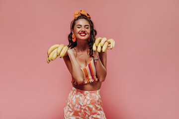 Wall Mural - Bright stylish woman with stylish accessories and red lips in striped colorful top and modern pink pants holding bananas and smiling with closed eyes..