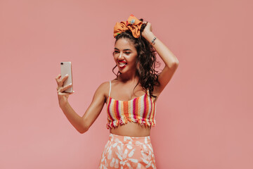 Wall Mural - Funny curly girl with stylish headband and accessories in colorful top and pineapple print pants makes photo on pink backdrop..