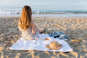 Wall Mural - Summer picnic on the beach at sunset. Young woman with glass of rose wine. Weekend picnic. Rear view, people from behind.