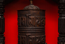 Tibetan Prayer Drum Made Of Copper On A Red Background. The Cylinder On The Axis Contains The Mantra. Image Of The Mantric Inscriptions Of Avalokiteshvara "om Mani Padme Hum" On The Prayer Drum.