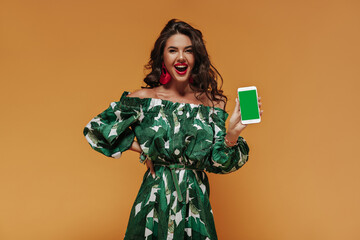 Positive curly haired woman with red lips and cool earrings in green modern sundress demonstrate white smartphone and looking into camera..