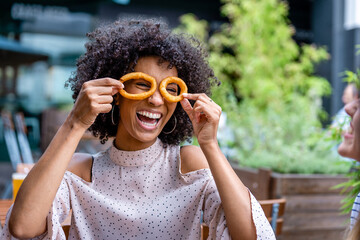 young and smiling African descending woman wearing fried onion rings as eyeglasses, funny and playful portrait of a mixed race person at restaurant having fun with friends