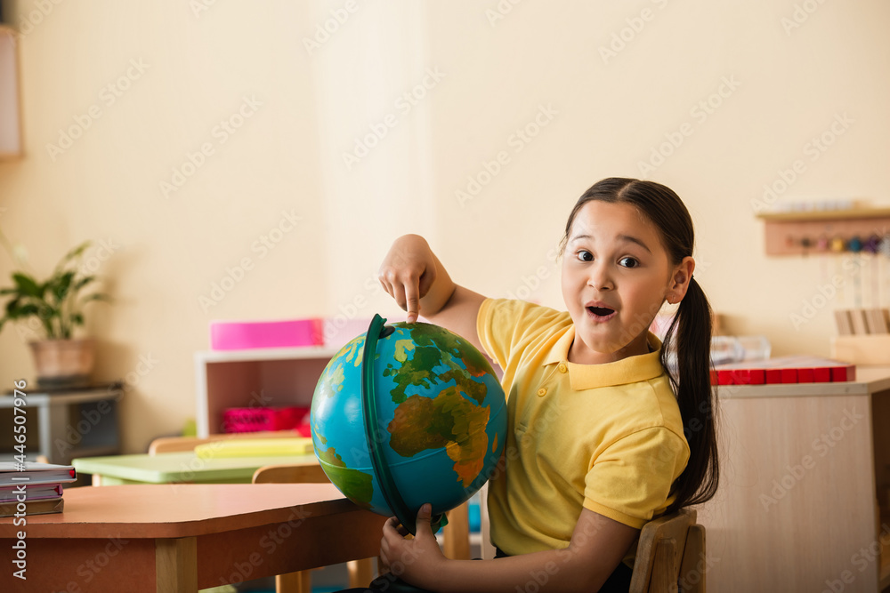 amazed asian girl pointing at globe and looking at camera in montessori school - obrazy, fototapety, plakaty 