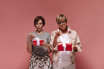 Wonderful two ladies with short hair in modern clothes and cool glasses holding red gift boxes and untying tapes on pink backdrop..