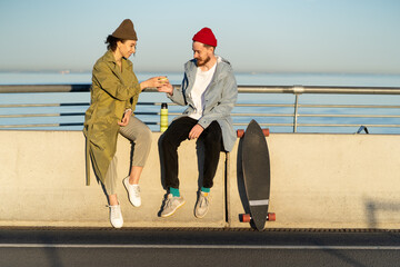 Happy couple drinking hot tea outdoors sitting on bridge broadwalk dressed in trendy street fashion clothes with longboard near. Young hipsters guy and girl refreshing after skateboard ride at sunset