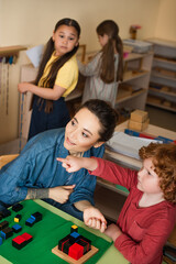 Wall Mural - boy pointing with finger while playing wooden cubes game near teacher and blurred interracial girls