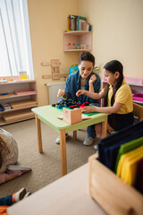 Wall Mural - asian girl playing wooden cubes game with teacher near kids on floor in classroom