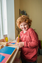 Wall Mural - cheerful, redhead boy looking at camera while holding color pencil in montessori school