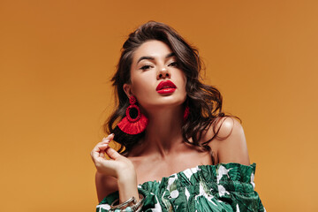 Trendy attractive woman with wavy hairstyle with modern cool earrings and big red lips in green sundress looking into camera..