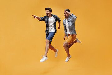 Wall Mural - Optimistic brunet young men in checkered shirts, white t-shirts and colorful shorts jump on orange background and point into distance.