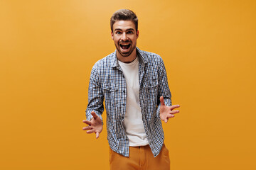 Wall Mural - Excited charming emotional man in blue checkered shirt and white t-shirt rejoices and looks into camera on orange background.