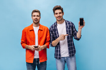 Wall Mural - Young handsome man in orange jacket smiles, brunet guy in checkered shirt point at phone screen on blue background.