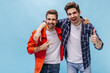 © Look! - Young brunet man in checkered shirt and handsome bearded guy in orange jacket rejoice and show peace sign and thumb up on blue background.
