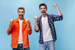 © Look! - Excited young bearded man in orange jacket and his friend in checkered shirt rejoice and smile on blue background.