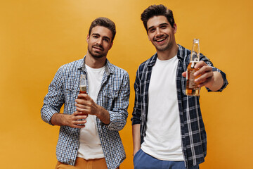 Handsome charming guys in stylish plaid shirts and white t-shirts pose on orange isolated background and hold beer bottles.