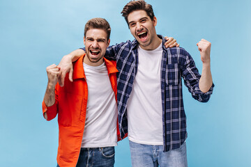 Wall Mural - Excited emotional men rejoice and scream on blue background. Charming man in checkered shirt and his friend in orange jacket pose on isolated.