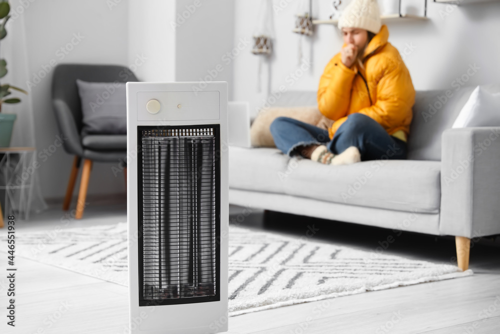 Young woman sitting in room with electric heater. Concept of heating season - obrazy, fototapety, plakaty 
