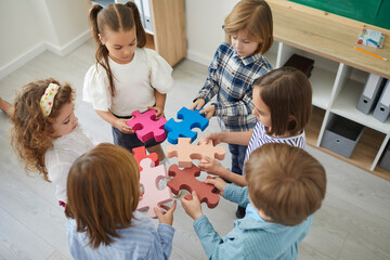 Wall Mural - Team of primary school children or kindergartners standing in circle join pieces of colorful jigsaw puzzle, high angle close up. Education, learning, engaging activities in classroom, teamwork concept