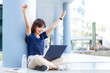 © kudosstudio - Happy young attractive asian business woman raises her arms up excitedly while looking at content on her laptop computer screen, sitting on the hallway floor