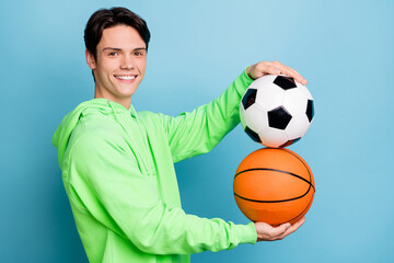 Photo of sweet cute young guy dressed green hoodie holding football basketball balls smiling isolated blue color background