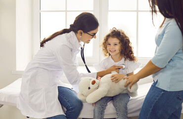 Wall Mural - Happy cute child patient with toy seeing family doctor. Female pediatrician with stethoscope checking young patient's heartbeat or doing her lungs exam during medical checkup at hospital or clinic