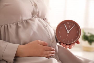 Wall Mural - Young pregnant woman holding clock near her belly at home, closeup. Time to give birth