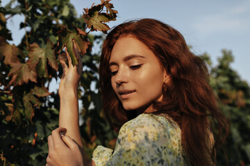 Wall Mural - Beautiful girl with foxy hair and freckles in yellow and green outfit looking down and posing on background of vineyards