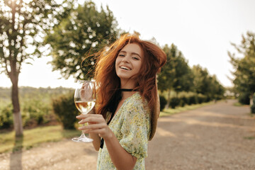 Wall Mural - Happy attractive girl with long ginger hair in summer trendy outfit looking into camera and holding glass of wine outdoor 