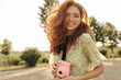 © Look! - Cheerful stylish lady with wavy long red hairstyle and cute freckles and straw hat smiling and holding pink camera outdoor..